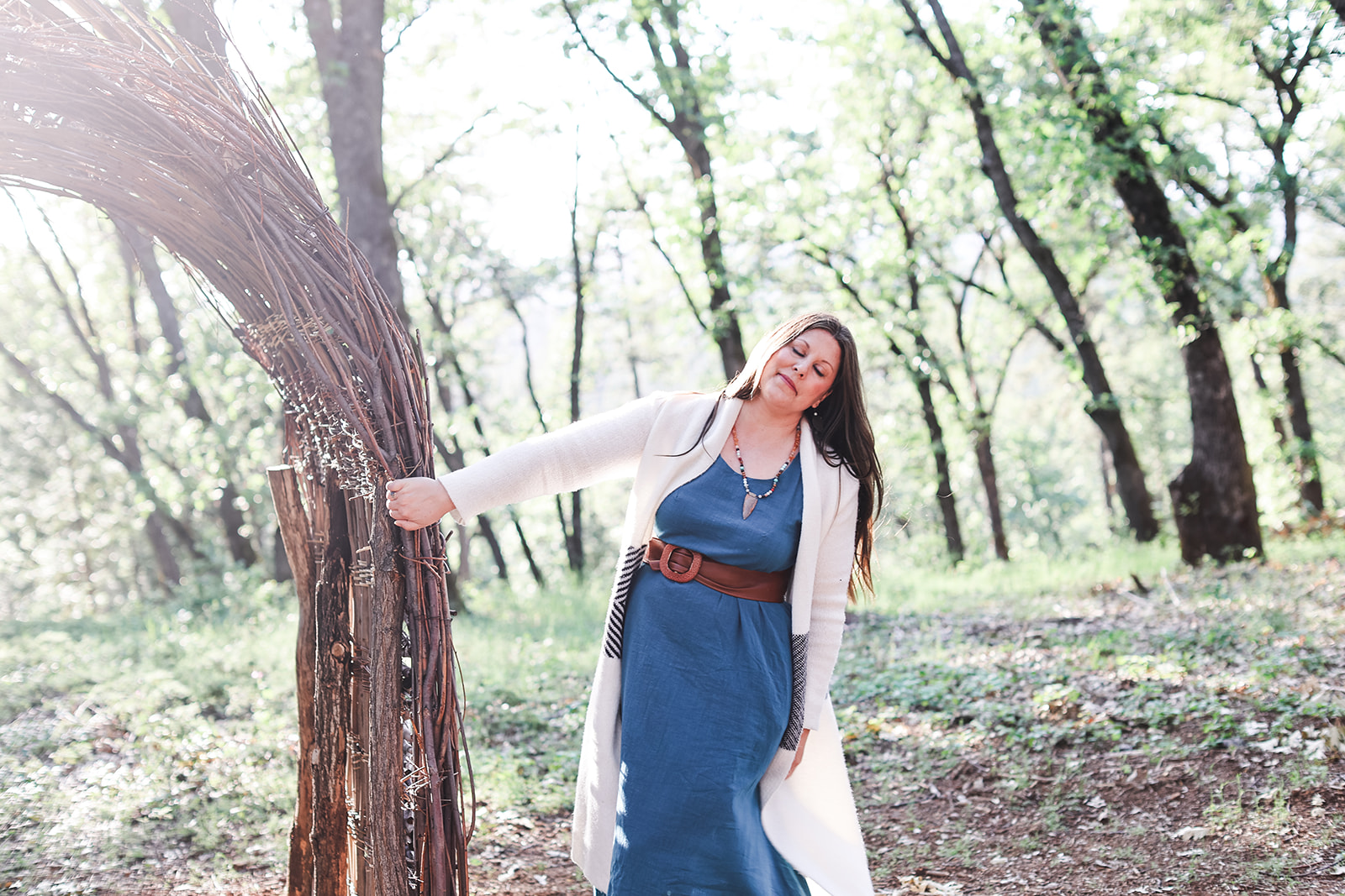 Amanda Smith stands in a sunlit forest, gently holding a handcrafted arch woven from natural branches.