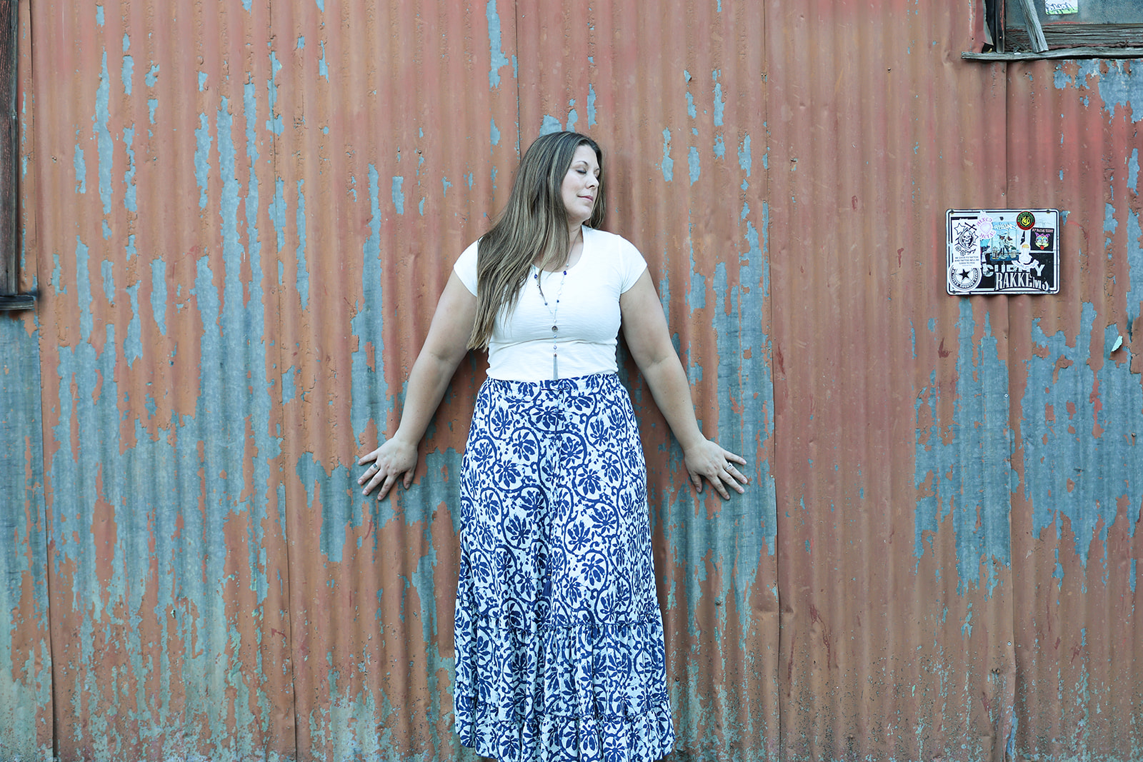 Amanda Smith standing against a weathered rust and blue metal wall, wearing a white top and blue floral maxi skirt.